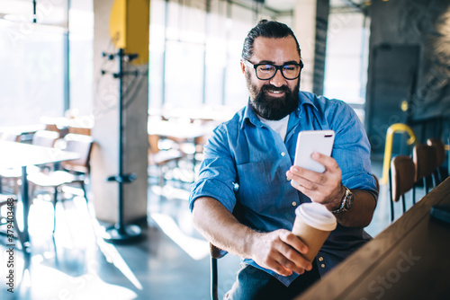 Happy confident mature male entrepreneur in casual wear sitting near bar station in cafe, cheerful hospitable bearded man owner of small business coffee shop checking notification on smartphone