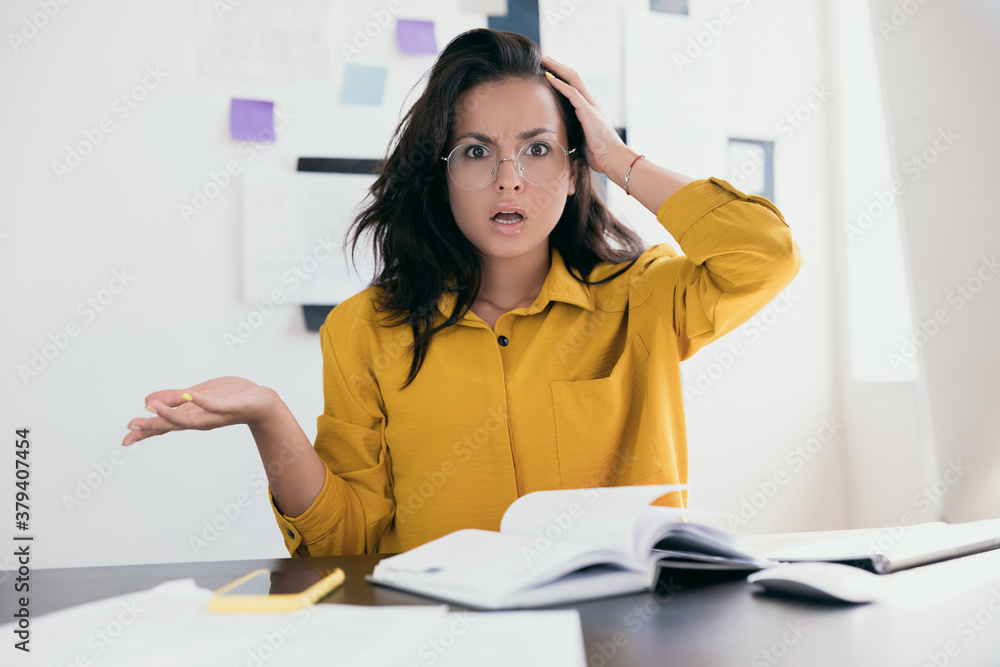 Unpleasantly surprised woman freelancer holding head. Lot of papers on desk. Notes and plans on background. Pretty women with brown hair wearing yellow shirt. Office work or work from home concept. Unpleasantly surprised woman freelancer holding head. Lot of papers on desk. Notes and plans on background. Pretty women with brown hair wearing yellow shirt. Office work or work from home concept.