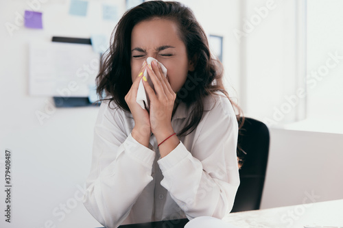 Sick and overworked businesswoman sitting at the desk in the office and blowing her nose  feeling unwell. Concept of hard work without maintaining health. Office work or work from home.