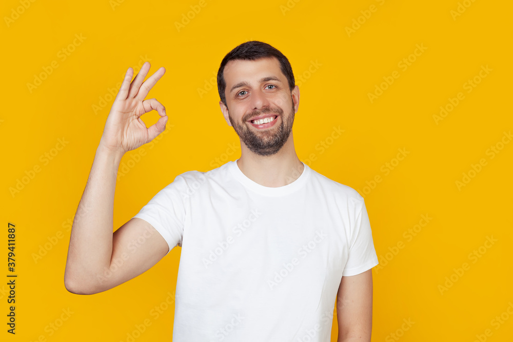 Young man with a beard in a white t-shirt happy face, smiling positively, makes okay sign with hand and fingers. It's okay. Lucky expression. Stands on isolated yellow background