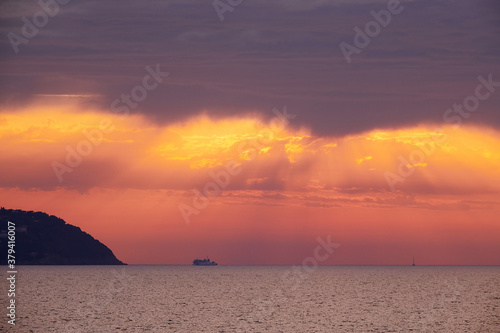 Mediterranean sea sunset during a storm. Orange sun rays filtered by the dark clouds. Tip of an island on the left side. A cruise boat navigates at the horizon.