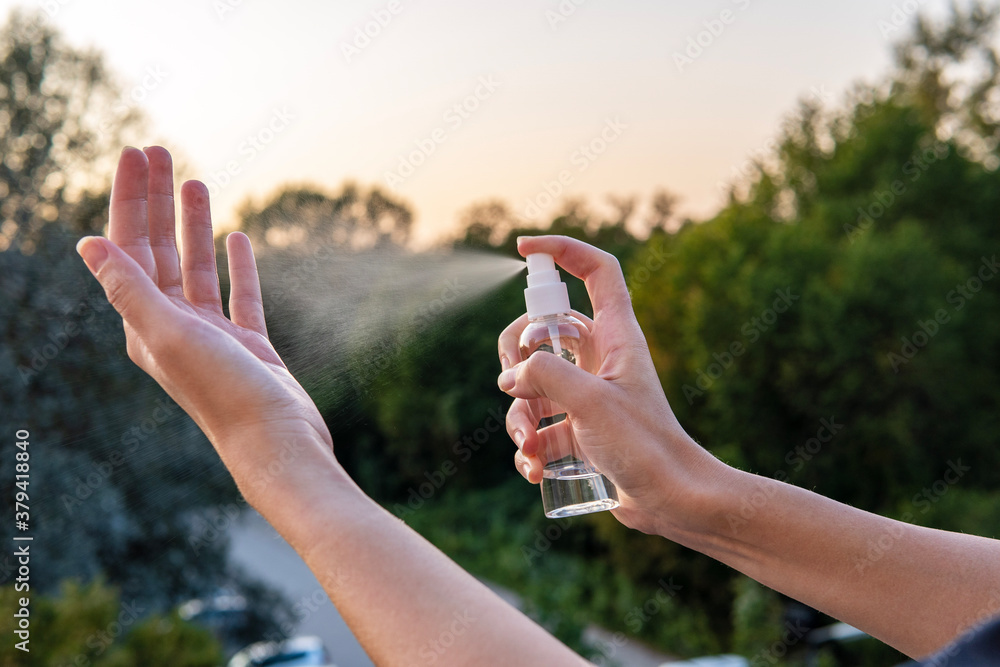 Woman use an alcohol hand sanitizer from a spray bottle to clean her hands and palms for protection from viruses, bacteria, and coronavirus. Beautiful golden sky in the background and green trees. - obrazy, fototapety, plakaty 