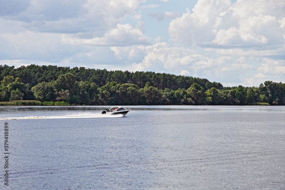 Obraz premium Fast floating motor boat on Russian Volga river with riverside forest and blue sky with clouds background, beautiful natural landscape at summer day, outdoor adventure, active recreation water tourism