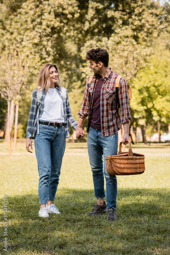 man with wicker basket holding hands with girlfriend while walking in park