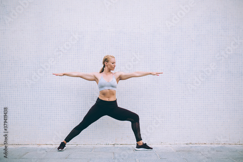 Energetic slim woman in tracksuit doing Warrior one during yoga exercises in morning, female gymnastic enjoying virabhadrasana during time for physical recreation at publicity area with copy space