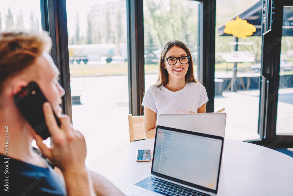 Portrait of cheerful woman in optical eyewear smiling at camera during cooperation brainstorming meeting with blurred male colleague on frontage using smartphone application for wireless calling