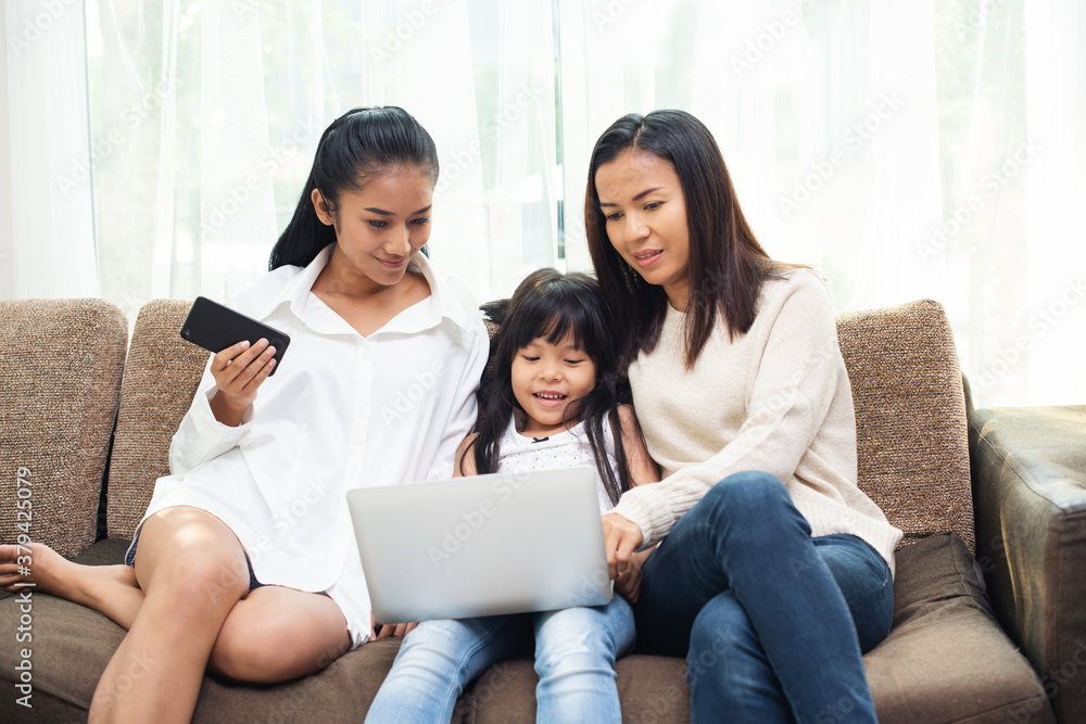 Happy together. Asian mother or female and her friend with child girl sitting on sofa watching fun movie on laptop, playing education game, studying language course for children Happy together. Asian mother or female and her friend with child girl sitting on sofa watching fun movie on laptop, playing education game, studying language course for children