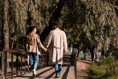 couple in trench coats holding hands and walking on wooden bridge in autumnal park