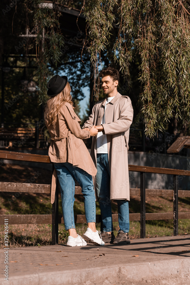 joyful man in trench coat looking at woman in hat while holding hands in park