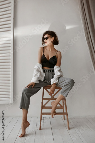 A model girl with perfect body in a black top and grey pants looking aside and posing on a stool indoors