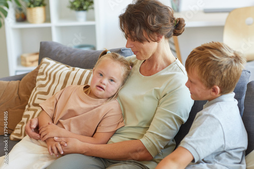 Portrait of modern mother embracing girl with down syndrome while sitting on couch with two kids in home interior