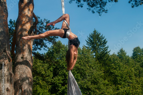 A slender girl with blonde hair, in a black stage costume, performs gymnastic and circus exercises on white silk against the background of the forest and the sky.