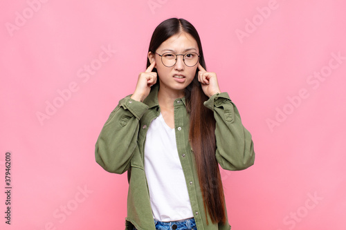 asian young woman looking angry, stressed and annoyed, covering both ears to a deafening noise, sound or loud music
