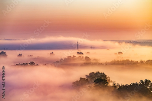 Beautiful panoramic landscape with river valley covered by thick fog in autumn in the early morning. Sunrise. Sun rays shine through the thick fog. Electric power transmission line in fog.