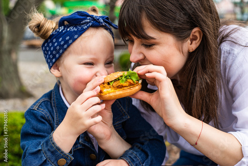 Young mother and little daughter ear burger together in outdoors cafe