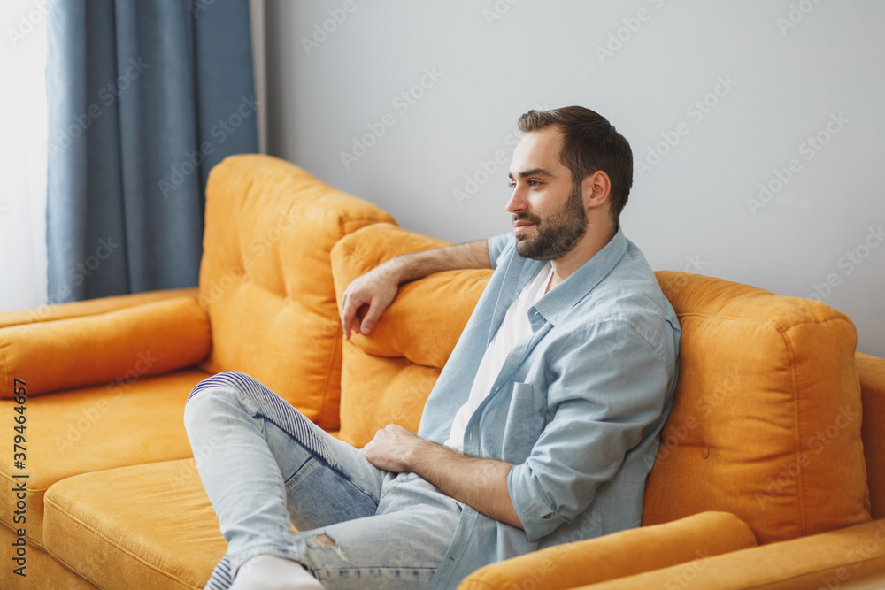 Φωτογραφία Smiling handsome attractive young bearded man 20s wearing casual white t-shirt blue shirt looking aside sitting on couch resting relaxing spending time in living room at home