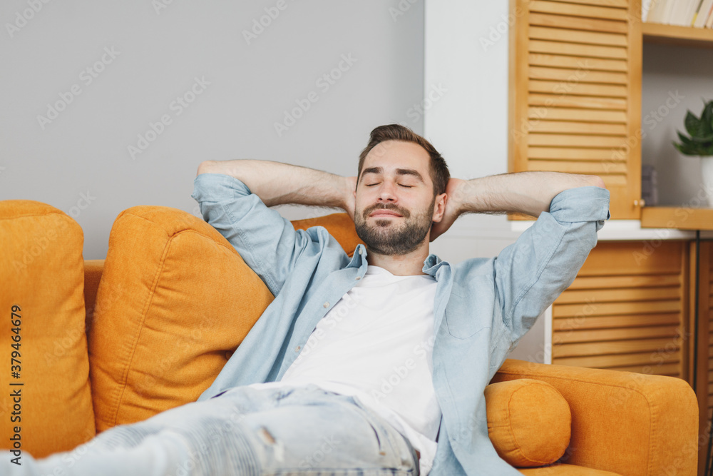 Φωτογραφία Attractive handsome young bearded man 20s wearing casual white t-shirt blue shirt looking aside sleeping with hands behind head lying on couch resting relaxing spending time in living room at home