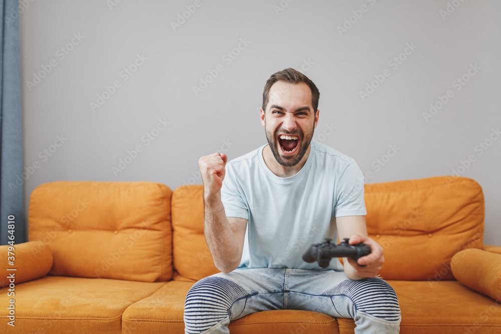 Crazy screaming cheerful young bearded man 20s wearing casual blue t-shirt play pc game with joystick console clenching fists sitting on couch resting relaxing spending time in living room at home. Crazy screaming cheerful young bearded man 20s wearing casual blue t-shirt play pc game with joystick console clenching fists sitting on couch resting relaxing spending time in living room at home.