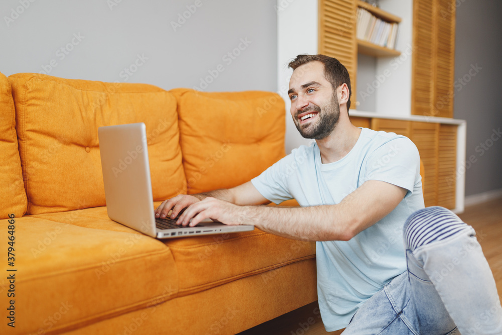 Smiling funny attractive handsome young bearded man 20s in casual blue t-shirt working on laptop pc computer sitting at floor near couch resting relaxing spending time in living room at home. Smiling funny attractive handsome young bearded man 20s in casual blue t-shirt working on laptop pc computer sitting at floor near couch resting relaxing spending time in living room at home.