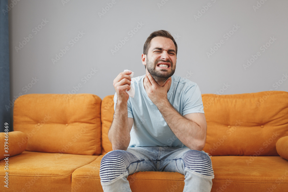 Sick exhausted young bearded man in casual blue t-shirt hold paper napkin coughing sneezing put hand on neck throat keeping eyes closed sitting on couch resting spending time in living room at home. Sick exhausted young bearded man in casual blue t-shirt hold paper napkin coughing sneezing put hand on neck throat keeping eyes closed sitting on couch resting spending time in living room at home.
