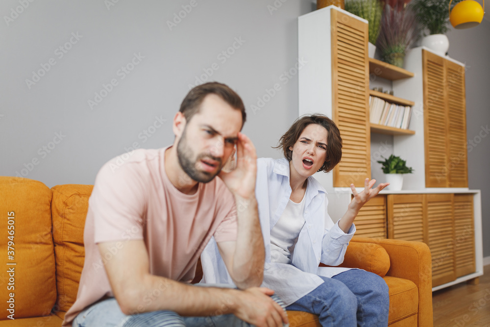 Displeased dissatisfied angry irritated young couple two friends man woman 20s in casual clothes sitting on couch screaming swearing spreading hands looking aside spending time in living room at home.