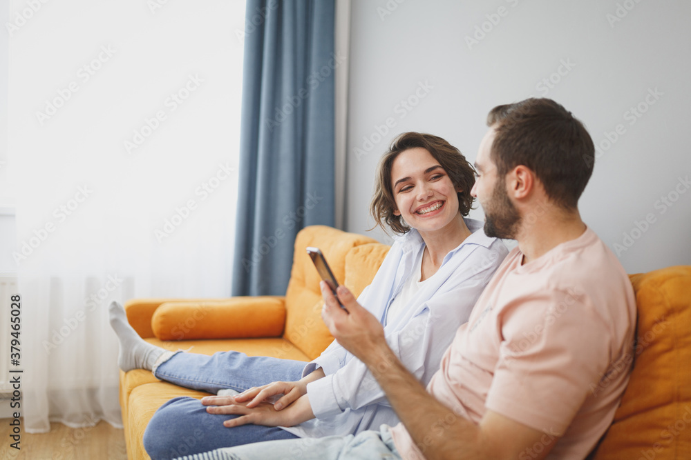 Funny smiling young couple two friends man woman 20s wearing casual clothes sit on couch hugging using mobile cell phone looking at each other resting relaxing spending time in living room at home. Funny smiling young couple two friends man woman 20s wearing casual clothes sit on couch hugging using mobile cell phone looking at each other resting relaxing spending time in living room at home.