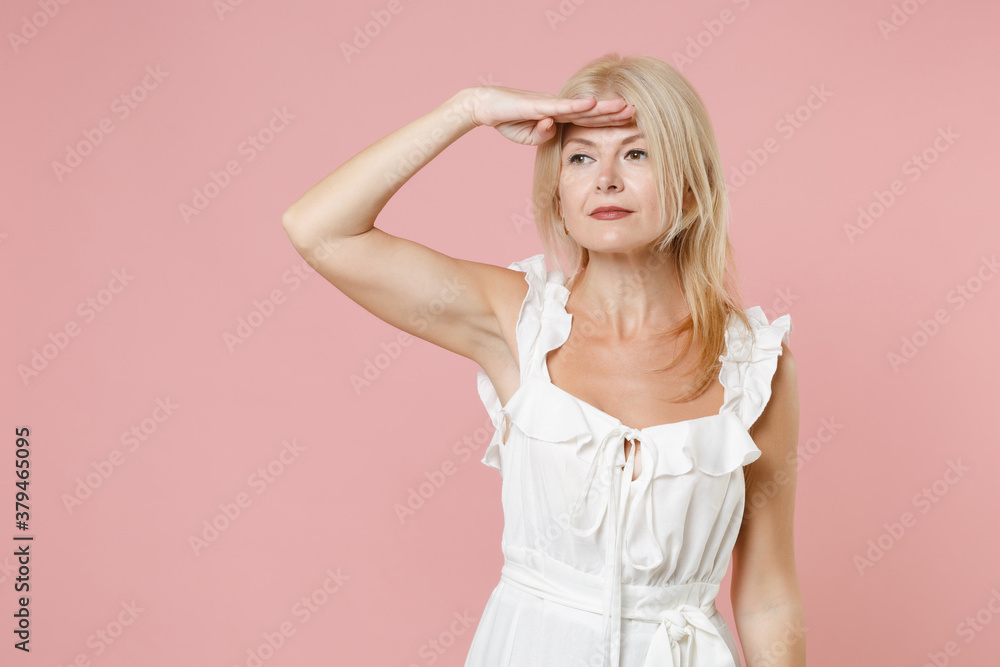 Smiling pretty beautiful attractive young blonde woman in white summer dress standing holding hand at forehead looking far away distance isolated on pastel pink colour background, studio portrait. Smiling pretty beautiful attractive young blonde woman in white summer dress standing holding hand at forehead looking far away distance isolated on pastel pink colour background, studio portrait.