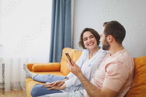 Funny smiling young couple two friends man woman 20s wearing casual clothes sit on couch hugging using mobile cell phone looking at each other resting relaxing spending time in living room at home.