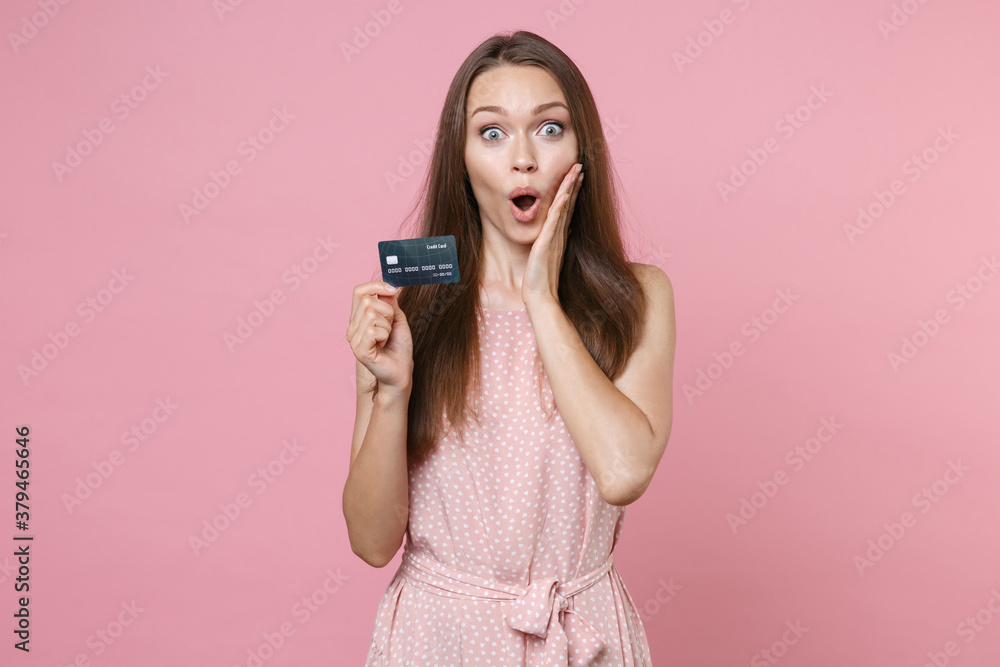 Shocked young brunette woman 20s wearing pink summer dotted dress posing holding in hand credit bank card put hand on cheek looking camera isolated on pastel pink color background studio portrait. Shocked young brunette woman 20s wearing pink summer dotted dress posing holding in hand credit bank card put hand on cheek looking camera isolated on pastel pink color background studio portrait.