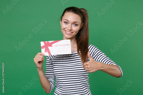 Smiling attractive young brunette woman 20s in striped casual clothes posing holding in hand gift certificate showing thumb up looking camera isolated on green color wall background studio portrait.