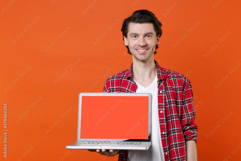 Smiling young man 20s in white t-shirt red checkered shirt posing hold laptop pc computer with blank empty screen mock up copy space looking camera isolated on orange wall background studio portrait. Smiling young man 20s in white t-shirt red checkered shirt posing hold laptop pc computer with blank empty screen mock up copy space looking camera isolated on orange wall background studio portrait.
