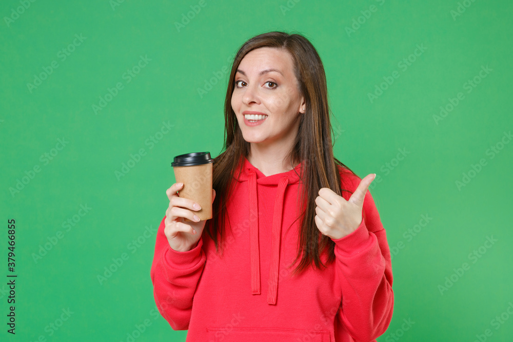 Fototapeta premium Smiling beautiful cheerful young brunette woman 20s wearing red casual streetwear hoodie posing showing thumb up hold paper cup of coffee or tea isolated on green color background studio portrait.