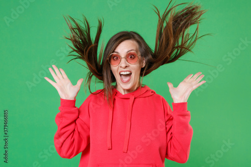 Excited surprised young brunette woman 20s in bright red casual streetwear hoodie eyeglasses posing keeping mouth open throwing fluttering hair isolated on green color wall background studio portrait.