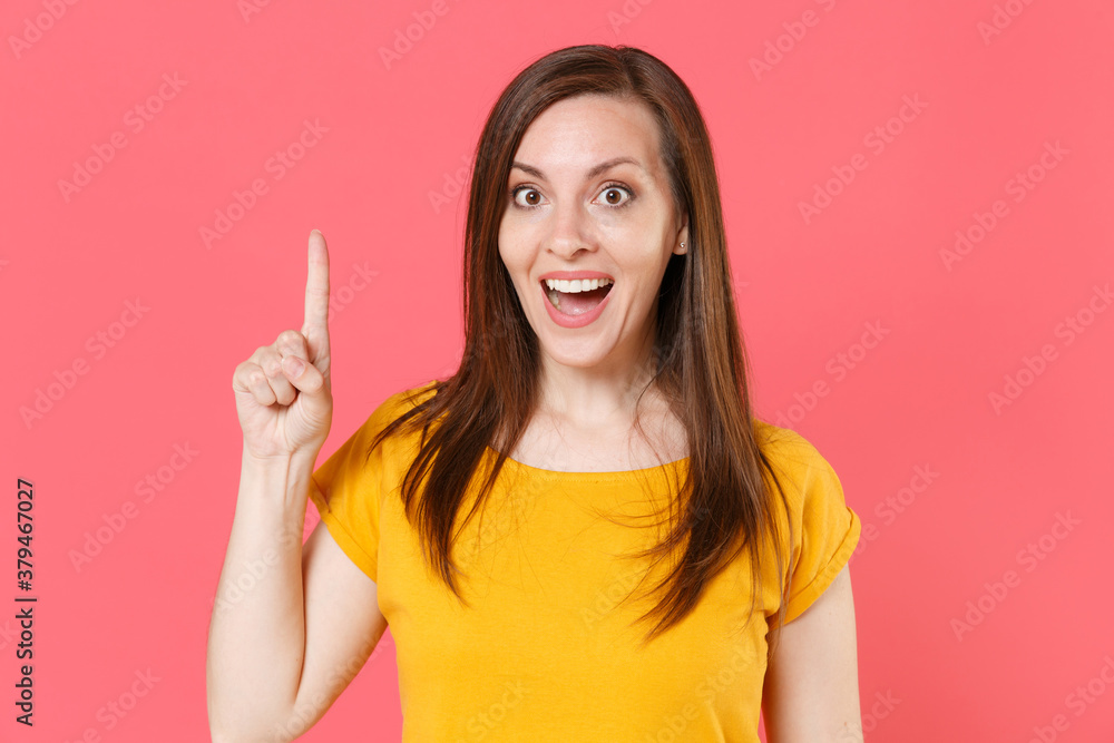 Excited shocked young brunette woman 20s wearing yellow casual t-shirt posing standing holding index finger up with great new idea looking camera isolated on pink color background studio portrait.