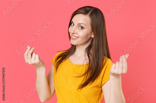 Smiling young brunette woman 20s wearing yellow casual t-shirt posing standing rubbing fingers showing cash gesture asking for money looking camera isolated on pink color background studio portrait.