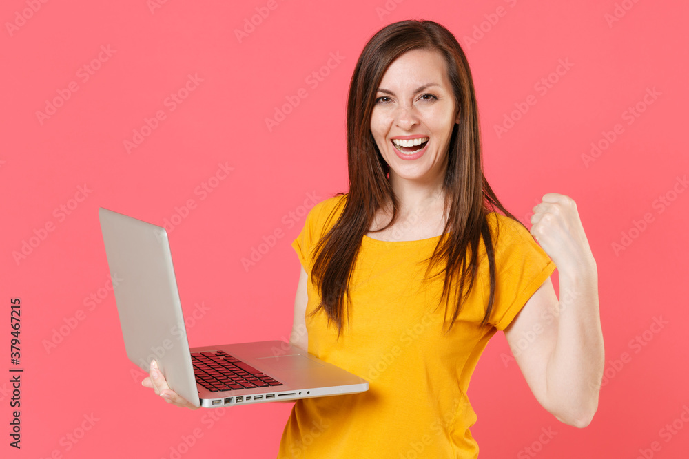 Naklejka premium Happy joyful young brunette woman 20s wearing yellow casual t-shirt posing and working on laptop pc computer doing winner gesture looking camera isolated on pink color wall background studio portrait.