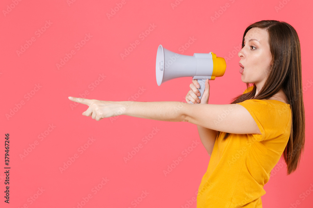 Side view of shocked young brunette woman in yellow casual t-shirt posing screaming in megaphone pointing index finger aside on mock up copy space isolated on pink color background studio portrait. Side view of shocked young brunette woman in yellow casual t-shirt posing screaming in megaphone pointing index finger aside on mock up copy space isolated on pink color background studio portrait.