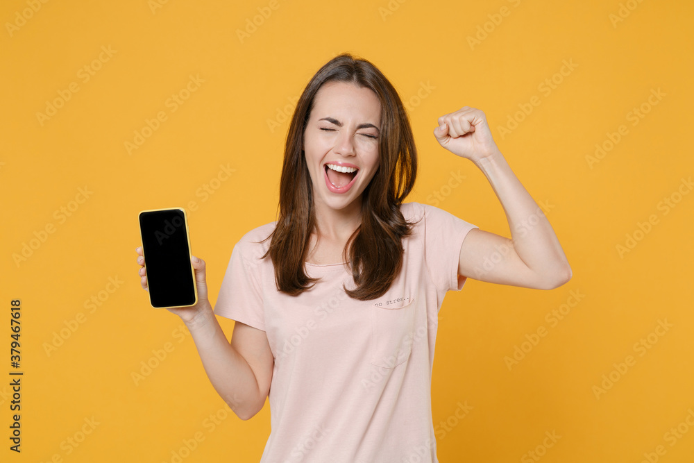 Happy young brunette woman in pastel pink casual t-shirt posing hold mobile cell phone with blank empty screen mock up copy space doing winner gesture isolated on yellow background studio portrait. Happy young brunette woman in pastel pink casual t-shirt posing hold mobile cell phone with blank empty screen mock up copy space doing winner gesture isolated on yellow background studio portrait.