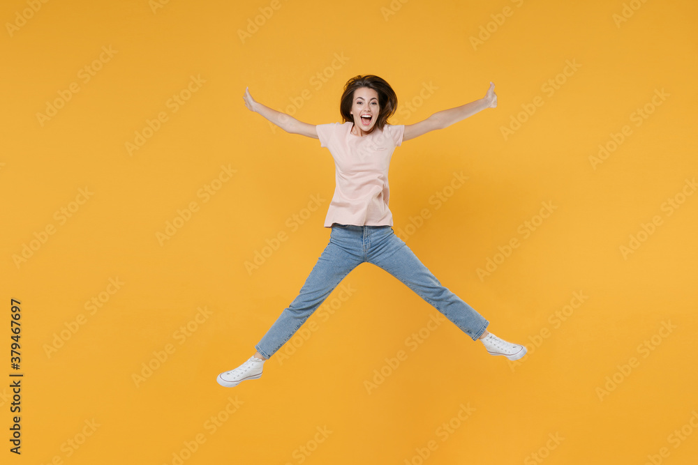 Full length portrait of excited happy young woman 20s wearing pastel pink casual t-shirt posing jumping spreading legs and hands looking camera isolated on bright yellow color wall background studio.