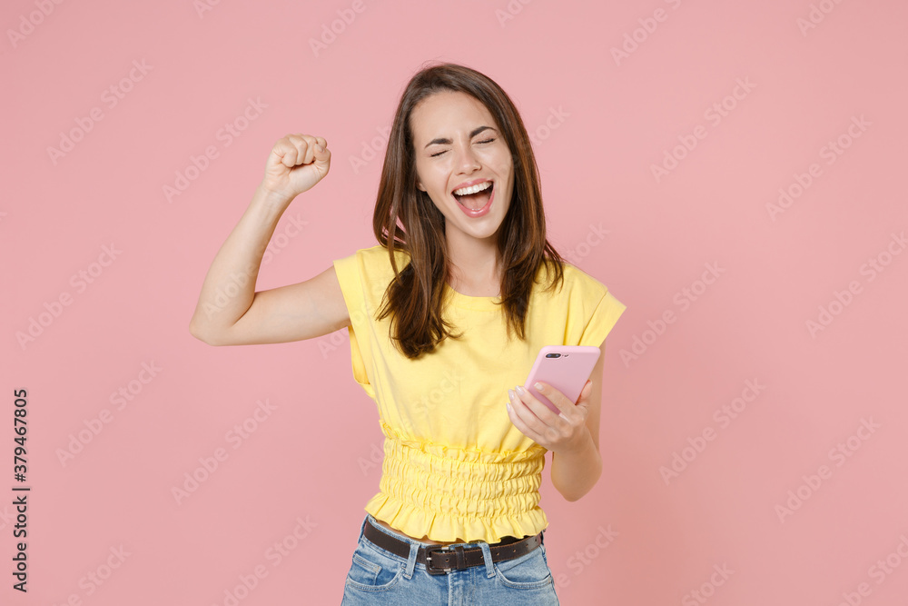 Happy young brunette woman 20s wearing yellow casual t-shirt posing standing using mobile cell phone typing sms message doing winner gesture isolated on pastel pink color background studio portrait.