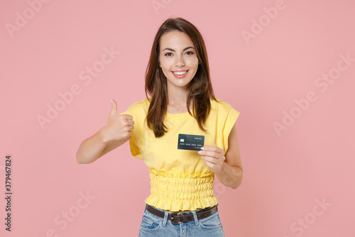 Smiling beautiful attractive young brunette woman in yellow casual t-shirt posing standing holding credit bank card showing thumb up looking camera isolated on pastel pink background studio portrait.