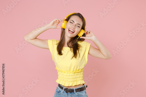Cheerful young brunette woman 20s wearing yellow casual t-shirt posing listening music with headphones put hands on head keeping eyes closed isolated on pastel pink color background studio portrait.