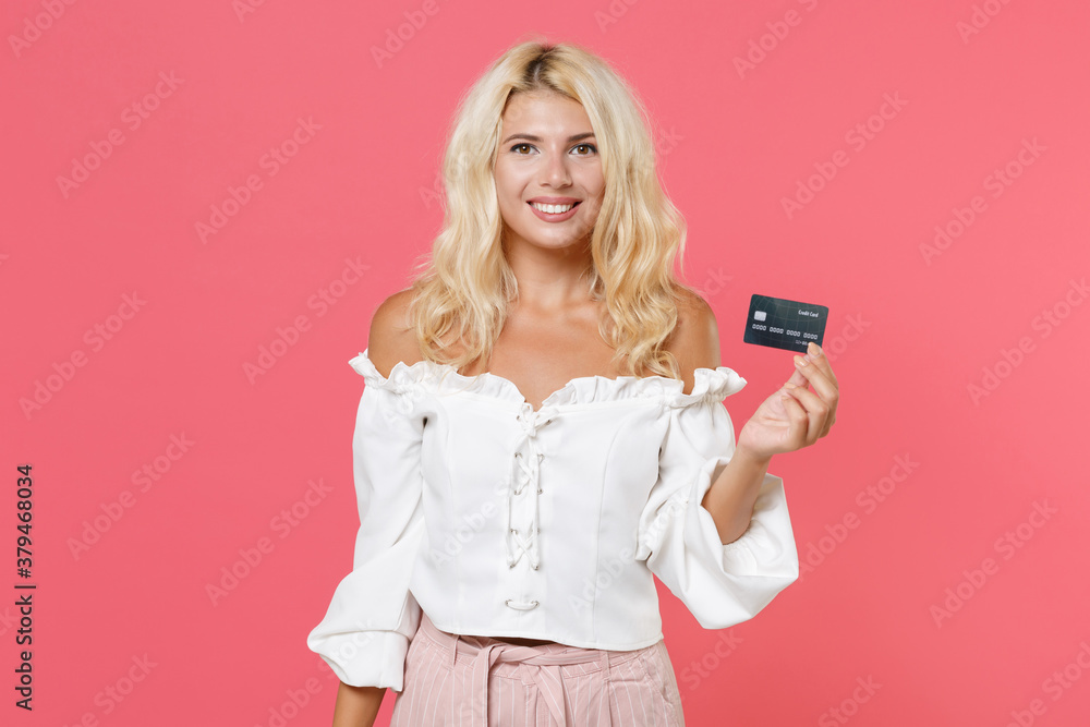 Smiling charming beautiful attractive young blonde woman 20s wearing white casual clothes holding in hand credit bank card looking camera isolated on bright pink colour background, studio portrait.