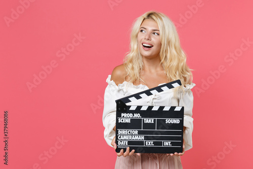 Excited cheerful young blonde woman 20s wearing white casual clothes standing hold classic black film making clapperboard looking aside isolated on bright pink colour background, studio portrait.