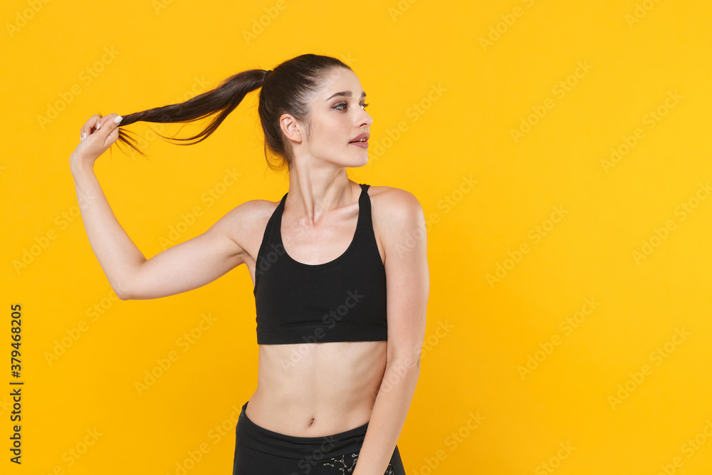 Beautiful attractive young fitness sporty woman 20s wearing black sportswear posing working out training holding hair ponytail looking aside isolated on bright yellow color background studio portrait. Beautiful attractive young fitness sporty woman 20s wearing black sportswear posing working out training holding hair ponytail looking aside isolated on bright yellow color background studio portrait.