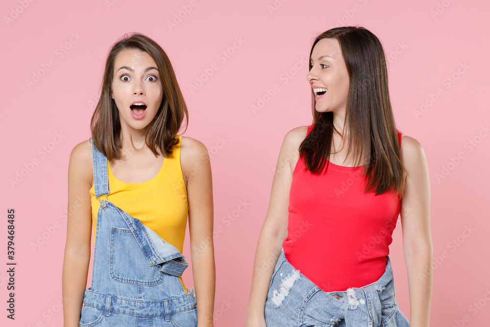 Two excited surprised shocked amazed attractive young women friends 20s in casual denim clothes keeping mouth open looking camera aside isolated on pastel pink colour background, studio portrait.