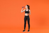 Full length portrait of attractive tired young fitness sporty woman in black sportswear posing training working out drinking water from bottle looking aside isolated on orange color background studio.