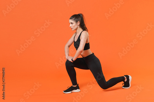 Full length side view portrait of beautiful young fitness sporty woman in black sportswear posing training working out doing exercise lunge looking aside isolated on orange color background studio.