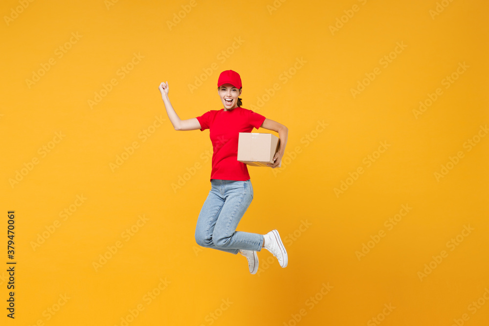 Full length body jumping delivery employee woman in red cap blank t-shirt uniform work courier in service during quarantine coronavirus covid-19 virus hold cardboard box isolated on yellow background.