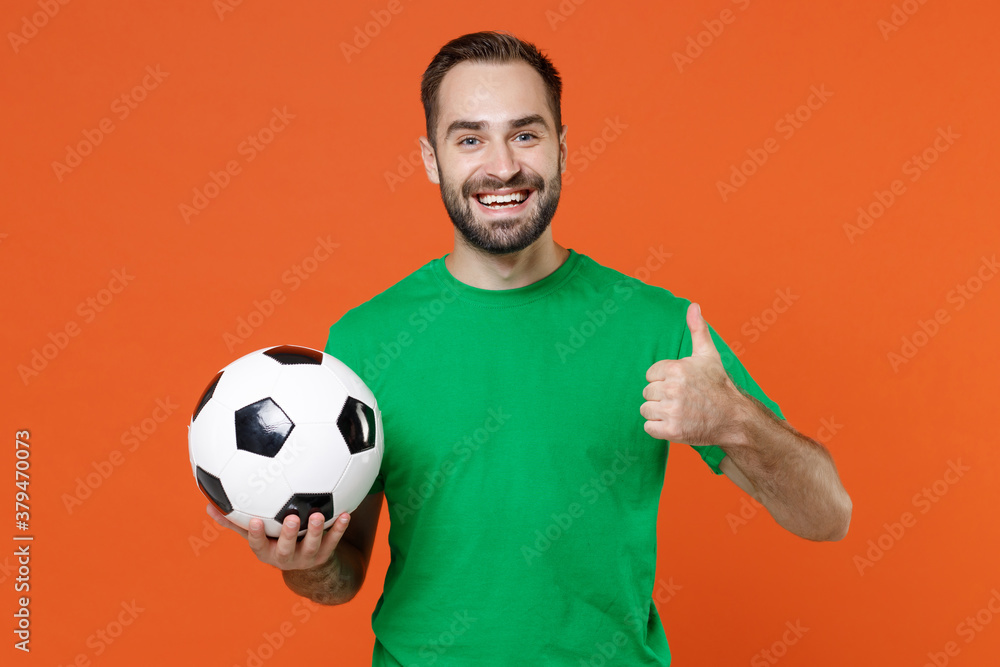Smiling young man football fan in green t-shirt cheer up support favorite team with soccer ball showing thumb up isolated on orange background studio portrait. People sport leisure lifestyle concept. Smiling young man football fan in green t-shirt cheer up support favorite team with soccer ball showing thumb up isolated on orange background studio portrait. People sport leisure lifestyle concept.